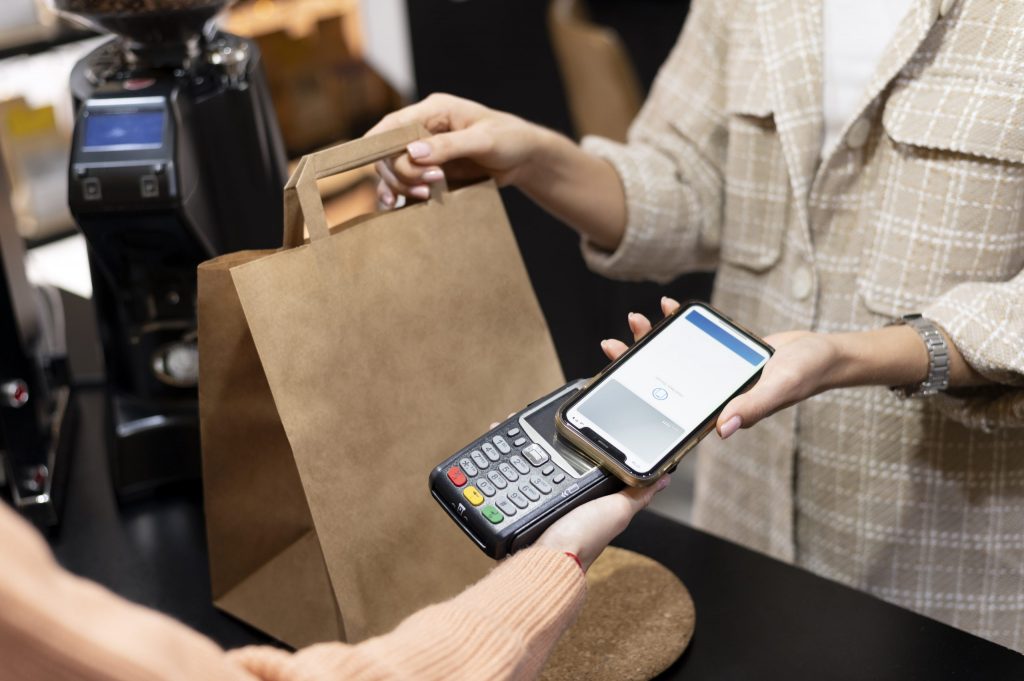 A person makes a contactless payment with a smartphone at a counter, while another person hands them a brown paper bag.